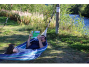 Person liegt in einer Hängematte und hält ein geöffnetes Buch neben einem Fluss