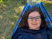 Person mit dunklen, lockigen Haaren und Brille liegt in einer blauen Hängematte auf Gras, trägt ein dunkles Oberteil