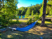 Blaue, gestreifte Hängematte zwischen zwei Bäumen über einer grasigen Fläche, im Hintergrund ein See und bewaldetes Ufer unter blauem Himmel