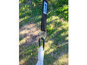 Nahaufnahme eines schwarzen Stoffgurts mit Etikett, befestigt an zwei Metallringen und einem Karabiner, an dem ein weißes geflochtenes Seil hängt; Gras im Hintergrund