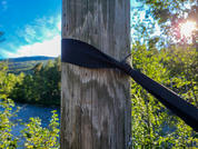 Nahaufnahme eines senkrechten Holzpfahls mit schwarzem Band; im Hintergrund Fluss, Bäume, Hügel und blauer Himmel mit Sonne