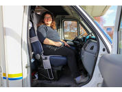 Person sitzt auf dem Fahrersitz eines weißen Transporters mit geöffneter Tür, trägt Brille, dunkelblaues Oberteil und schwarze Hose