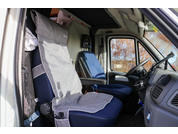 Innenraum einer Fahrzeugkabine mit zwei blauen Sitzen und grauen Sitzbezügen, Armlehnen, Lenkrad, Armaturenbrett und seitlichem Fenster mit Bäumen