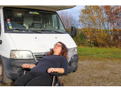 Person mit lockigem Haar und Brille sitzt im Rollstuhl vor einem weißen Transporter auf einem Schotterplatz