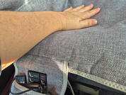 Hand und Unterarm auf grauem, strukturiertem Polsterstoff; unten links eine kleine schwarze Bedieneinheit mit Tasten und Kabel, daneben ein Reißverschluss und eine Öffnung mit schwarzer Unterlage
