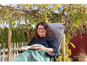 Person mit dunklen lockigen Haaren und Brille sitzt in einem Gartenstuhl, hält ein aufgeschlagenes Buch im Schoß, lächelt; Baum mit gelben Blättern darüber, Zaun und rotes Gebäude im Hintergrund