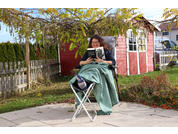 Person sitzt auf einer Gartenliege unter Weinlaub, liest ein Buch, grüne Decke über den Beinen, kleiner Klapptisch mit Thermosflasche und Tasse, roter Geräteschuppen im Hintergrund, Holzzaun und Rasen