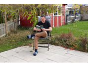 Person sitzt auf einem Gartenstuhl auf gepflasterter Fläche und hält ein aufgeschlagenes Buch; im Hintergrund Gartenhaus, Holzzaun, Rasen und Sträucher