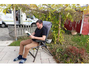 Eine Person sitzt auf einem Klappstuhl im Garten und liest ein aufgeschlagenes Buch; im Hintergrund Wohnmobil, roter Geräteschuppen, Bäume und Pflanzen