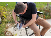 Person mit kurzem Haar, schwarzem Shirt und braunen Hosen sitzt auf einem Stuhl im Freien und hält ein graues rechteckiges Objekt; Rasen, Pflanzen und Laub sichtbar