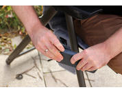 Zwei Hände halten ein dunkelgraues Smartphone und eine schwarze Klemmhalterung an einem Metallbein eines Klappstuhls. Sichtbarer Ring am linken Ringfinger, braune Shorts, Betonboden mit trockenen Blättern und einem Zweig