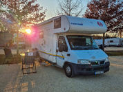 Weißes Wohnmobil auf einem Kiesstellplatz bei Sonnenuntergang, links Klappstühle und ein Tisch mit zwei Personen, Bäume im Hintergrund