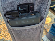 Graue Stoffseitentasche an einem Sitz mit zwei schwarzen Elektronikgeräten; oben kleines Bedienfeld mit den Beschriftungen 'Upper' und 'Lower' und angeschlossenem Kabel, darunter längliches Gerät mit USB-Anschluss und vier weißen Symbolen; im Hinterg