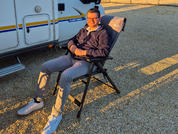 Person sitzt auf klappbarem Campingstuhl neben einem Wohnmobil auf kiesigem Boden, trägt dunkle Jacke, helle Hose und weiße Schuhe; tief stehende Sonne wirft lange Schatten