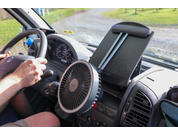 Autoinnenraum mit Lenkrad und Händen am Lenkrad, runder Ventilator an der Mittelkonsole, Tablet‑Halterung auf dem Armaturenbrett, Blick durch die Windschutzscheibe auf einen Weg und Gras