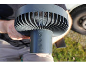 Hand hält einen runden Ventilator, Blick auf Lüftergitter, Bedientasten und LED-Punkte; im Hintergrund Gras und ein Reifen