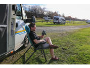 Person sitzt auf Klappstuhl neben weißem Van mit offener Tür und hält ein Lenkrad; weitere Vans auf grasigem Parkplatz im Hintergrund