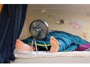 Füße ragen aus einem Schlafsack; ein kleiner Ventilator steht davor, im Hintergrund eine Wand mit mehreren Zeichnungen