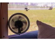 Kleiner runder Ventilator auf einer Ablage vor einem Fenster, rechts ein Kissen, draußen unscharfe Wiese