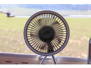 Runder Tischventilator mit Schutzgitter auf Dreibeinständer auf Fensterbank, im Hintergrund unscharfer Außenbereich mit Gras