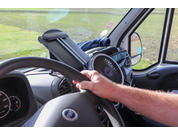 Hand und Unterarm an einem Lenkrad im Fahrzeuginneren; Armaturenbrett mit kleinem Ventilator und Halterung, Außenspiegel und Wiese durch die Windschutzscheibe sichtbar