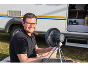 Mann mit Brille sitzt vor einem Wohnmobil und hält einen kleinen Ventilator auf einem Stativ; offenes Fach mit Schuhen und Flasche sichtbar