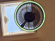 Runder Ventilator mit Schutzgitter und hellem grünen Leuchtring vor geöffnetem rechteckigem Dachfenster, blauer Himmel sichtbar