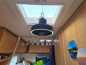 Innenraum mit offenem Dachfenster, hängende runde schwarze Lampe mit hellem Leuchtring, Reihe kleiner Glühbirnen an einer Leiste, Holzwände und Schränke, Bett mit grüner und blauer Decke und Kissen, dunkler Vorhang, zwei Landschaftsfotos an der Wand