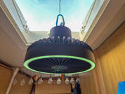 Runde schwarze Hängeleuchte mit grün leuchtendem Ring und zentralem Gitter, an Kabel und Haken unter einem geöffneten Dachfenster; Holzverkleidung an den Wänden, eine Reihe freiliegender Glühbirnen dahinter, dunkles Kleidungsstück und blaues Objekt a