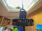 Schwarzes rundes Gerät mit senkrechtem Aufsatz, an einem blauen Karabinerhaken von der Decke vor einem offenen Dachfenster hängend; im Hintergrund Holzvertäfelung, Lichterkette und bunte Bettdecke