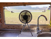 Kleiner runder Ventilator auf dreibeinigem Ständer auf einer Küchentheke vor einem rechteckigen Fenster; rechts Spüle mit Wasserhahn; draußen Feld und Bäume