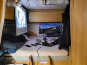 Innenraum mit Matratze, Kissen und Decken; links eine Leinwand mit projiziertem Bild; großes Foto an der Rückwand; Deckenlampen mit sichtbaren Glühbirnen; auf der Matratze Schüssel, Tasse, Kopfhörer, mehrere elektronische Geräte und Kabel; Leiter am 