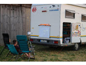 Wohnmobil von hinten auf einer Wiese, weißes quadratisches Paneel mit bunten Formen an der Heckklappe, zwei Klappstühle davor, eine Person sitzt auf einem Stuhl, Seitentür offen mit Blick ins Innere, orangerotes Kabel auf dem Boden, Holzsichtschutz i