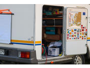 Geöffnete Seitentür eines weißen Wohnmobils mit Regalfächern, Kisten, aufgerollten Schläuchen und Kabeln; Innenseite der Tür mit Europa-Karte und Länderflaggenaufklebern