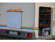 Heckansicht eines Wohnmobils mit heruntergelassener Leinwand, auf der ein unscharfes Bild sichtbar ist; geöffnete Seitentür mit Kisten, Kabeln und Werkzeug; gelb-blaue Streifen, Kennzeichen SIG SB 279