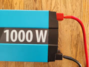 Blauer Wechselrichter mit großer weißer Beschriftung '1000 W', rote und schwarze Kabel, Holzuntergrund
