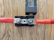 Zwei rote Kabel mit Metallmuttern an einem schwarzen Kunststoffhalter, dazwischen ein durchsichtiges Bauteil mit der Aufschrift "150", auf einer Holzoberfläche