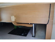 Unterseite einer Holzplatte, umwickeltes isoliertes Rohr mit weißem Winkel, schwarzes Kabel entlang der Kante, Metallfüße, schwarze Matte auf grauem Boden, teilweise sichtbarer grüner Plastikbeutel