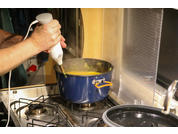 Hände halten einen weißen Stabmixer in einem blauen Kochtopf mit gelber Masse auf einem Gasherd, Dampf steigt auf