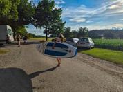 Persona con cabello largo llevando una tabla de stand-up paddle por una carretera rural; coches y una furgoneta aparcados, árboles, campo de maíz y cielo nublado