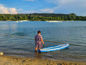 Persona está de pie con el agua hasta las rodillas en la orilla arenosa sosteniendo una tabla de paddle azul y blanca; obstáculos blancos en el lago, orilla arbolada y cielo azul con nubes al fondo