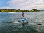 Persona de pie sobre una tabla de paddle azul y blanca con remo en un lago, boya con cuerda en primer plano, orilla con árboles y playa al fondo, cielo parcialmente nublado