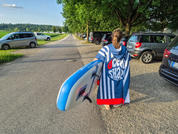Persona vista de espaldas que lleva un poncho a rayas azul y blanco y sostiene bajo el brazo una tabla de paddle surf azul y blanca, caminando por una carretera con coches aparcados, árboles y un prado bajo un cielo azul