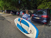Persona vista de espaldas llevando una tabla de paddle surf azul y blanca junto a coches aparcados y árboles