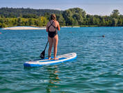 Persona con traje de baño negro de pie sobre una tabla de paddle surf azul y blanca en un lago tranquilo, sosteniendo un remo; orilla con árboles y una pequeña playa de arena bajo un cielo despejado al fondo