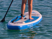 Pies y pantorrillas de una persona sobre una tabla de paddle azul en agua tranquila; remo visible