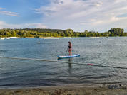 Persona en una tabla de paddle surf en un lago cerca de una orilla arenosa, cuerda a través del agua, línea de árboles y casas dispersas en la orilla opuesta, cielo azul con nubes