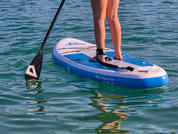 Piernas inferiores y pies de una persona sobre una tabla de paddle surf azul; remo tocando el agua verdeazulada