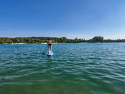 Persona de pie sobre una tabla de paddle en un lago, orilla arbolada en el fondo y cielo azul