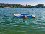 Persona apoyada en una tabla de paddle en un lago, espalda visible, agua verde, orilla arbolada y cielo azul al fondo